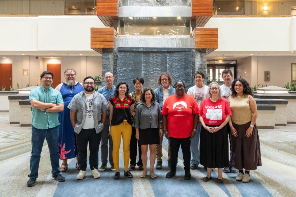 Thirteen people of varying heights, ages, and races stand in a hotel lobby with waterfall in background.